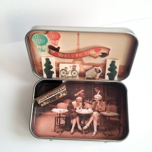 May include: A vintage-style tin box with a scene of two women sitting at a cafe table in Paris. The box has a red and white banner that says "J'aime Paris" and a small Eiffel Tower in the background. The box is open and the inside is decorated with a scene of a Parisian cafe.