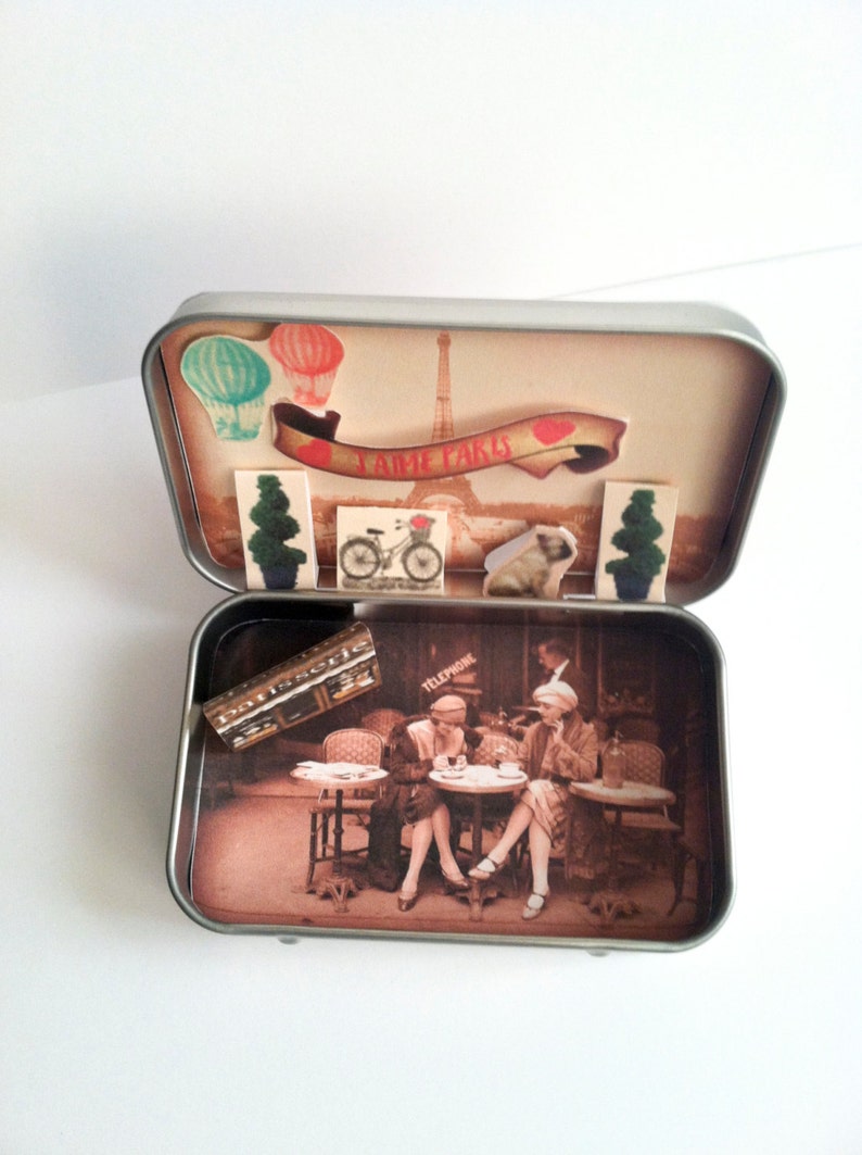 May include: A vintage-style tin box with a scene of two women sitting at a cafe table in Paris. The box has a red and white banner that says "J'aime Paris" and a small Eiffel Tower in the background. The box is open and the inside is decorated with a scene of a Parisian cafe.