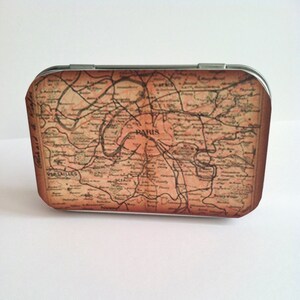 May include: A vintage-style metal tin with a map of Paris printed on the front. The map is in shades of brown and beige, with the word "PARIS" printed in the center.