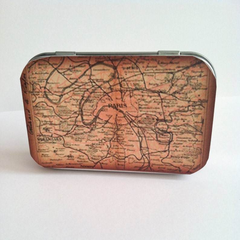 May include: A vintage-style metal tin with a map of Paris printed on the front. The map is in shades of brown and beige, with the word "PARIS" printed in the center.