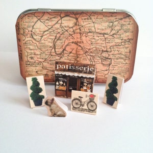 May include: A vintage tin box with a map of Paris on the lid. The box contains miniature figurines of a dog, a bicycle, two trees, and a patisserie.