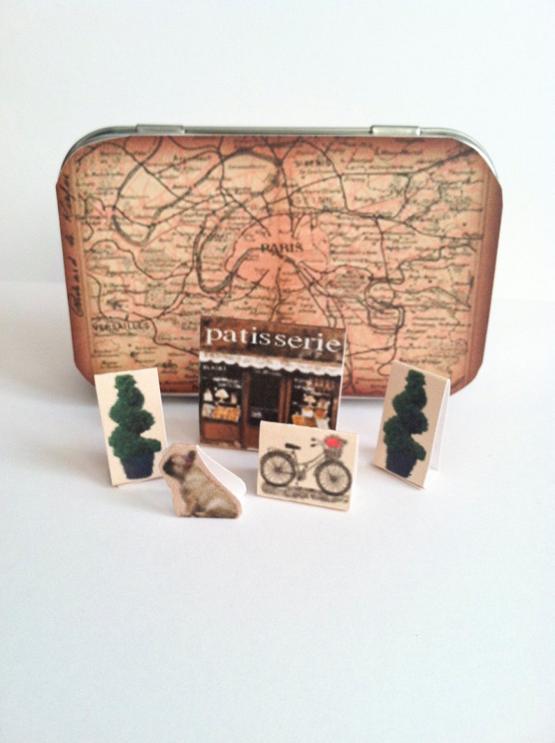 May include: A vintage tin box with a map of Paris on the lid. The box contains miniature figurines of a dog, a bicycle, two trees, and a patisserie.