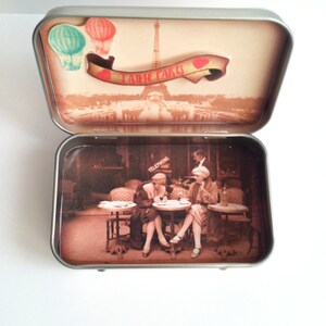 May include: A vintage-style tin box with a scene of the Eiffel Tower and the words "J'aime Paris" on the lid. The inside of the box features a sepia-toned image of three women sitting at a cafe table.
