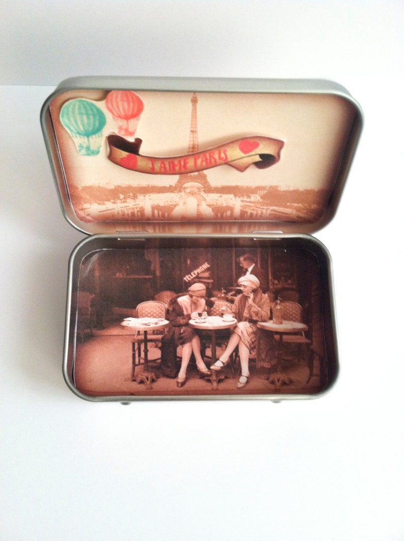 May include: A vintage-style tin box with a scene of the Eiffel Tower and the words "J'aime Paris" on the lid. The inside of the box features a sepia-toned image of three women sitting at a cafe table.