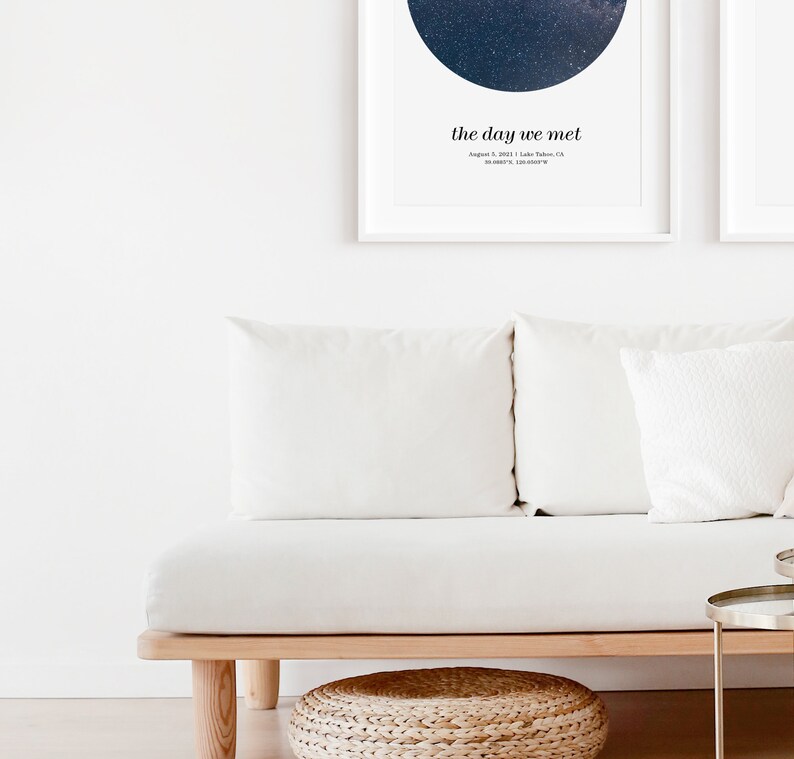 May include: Two framed prints with a starry night design and the text "the day we met" above a date and location. Below, a white sofa with white cushions and a woven footstool.