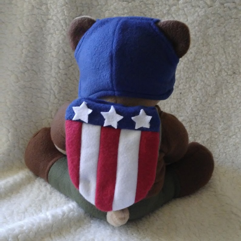 May include: A brown teddy bear wearing a blue and white Captain America costume. The costume includes a hat and a shield with stars.