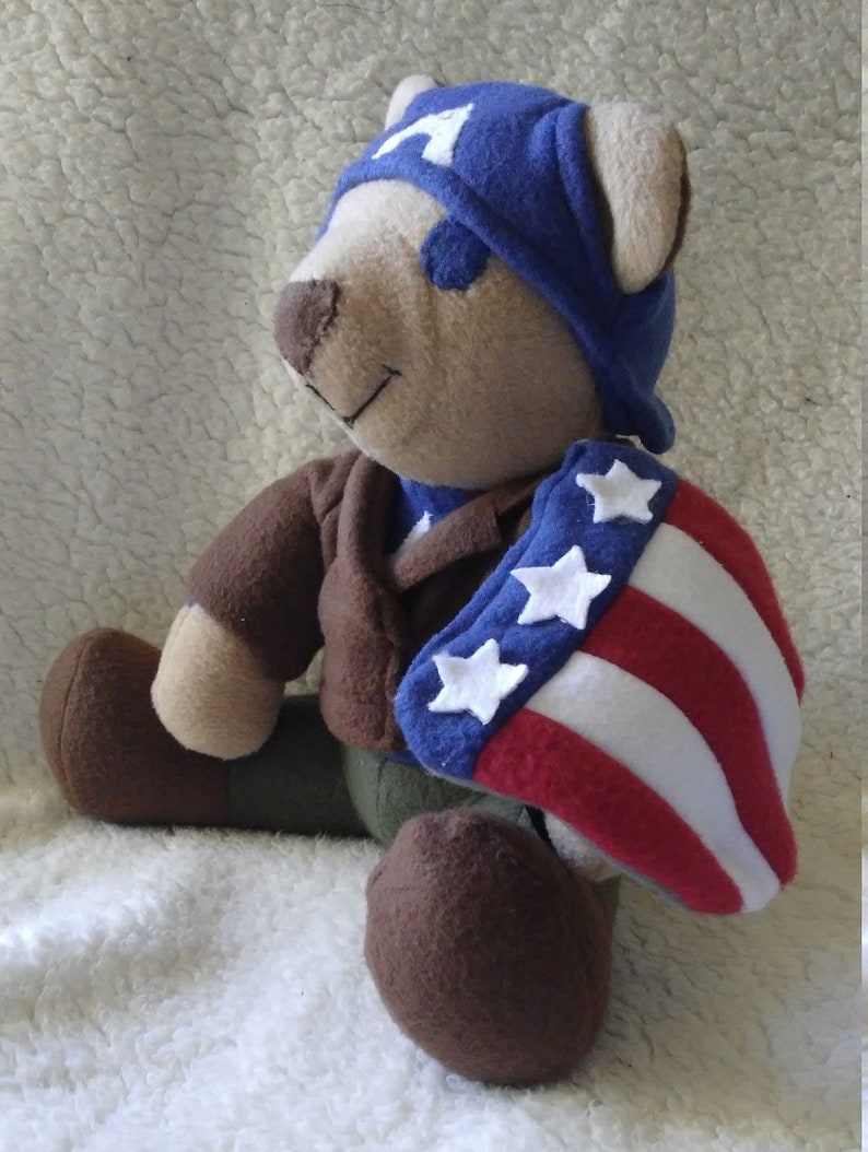 May include: A brown teddy bear wearing a blue and white cap and a red, white, and blue cape with white stars. The bear is sitting on a white fluffy surface.