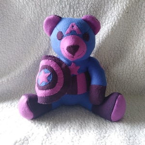 May include: A blue and purple teddy bear wearing a pink and purple Captain America costume. The bear is holding a shield in its left paw.