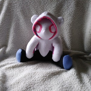 May include: A white plush toy in the shape of a bear wearing a hooded costume with pink and blue accents. The bear has black legs with blue feet. The hood has pink trim and blue stitching around the eye openings.