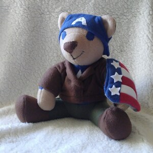 May include: A plush teddy bear dressed as Captain America. The bear is wearing a blue and red costume with a white star on the chest and a blue hat with a white "A" on the front. The bear is sitting on a white fluffy surface.