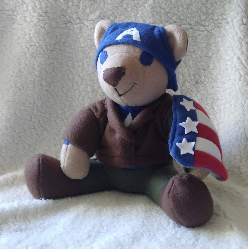 May include: A plush teddy bear dressed as Captain America. The bear is wearing a blue and red costume with a white star on the chest and a blue hat with a white "A" on the front. The bear is sitting on a white fluffy surface.