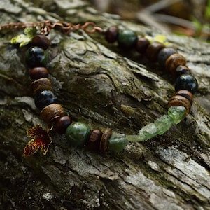 May include: A beaded bracelet featuring alternating dark brown wooden beads, smooth dark green spherical beads, and translucent light green raw crystal fragments. A small copper-toned maple leaf charm hangs from the side of the bracelet chain.