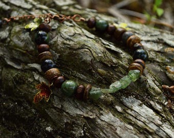 May include: A beaded bracelet featuring alternating dark brown wooden beads, smooth dark green spherical beads, and translucent light green raw crystal fragments. A small copper-toned maple leaf charm hangs from the side of the bracelet chain.