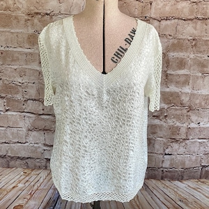 Puede incluir: Una blusa de manga corta color crema con cuerpo de tela texturizada y arrugada, escote en V con un borde acanalado ancho y detalles de malla de punto abierto en las mangas y el dobladillo inferior, sobre un maniquí.