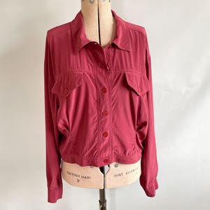 May include: A long-sleeved, red jacket with a collared neckline and two front pockets. The jacket has a button-up closure and is displayed on a vintage dress form. The fabric appears to be lightweight and slightly shiny.
