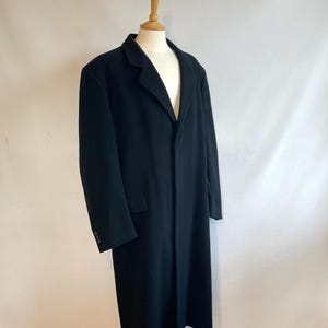 May include: A full-length black overcoat on a mannequin. The coat has a traditional notched lapel, a single-breasted closure, and a hem that reaches the knees. The sleeves have button details. The coat is likely made from a wool blend material.