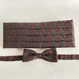 Op de afbeelding: Een formele set bestaande uit een vlinderdas en een cummerbund. De set heeft een patroon van rode bladeren op een donkergrijze achtergrond. De cummerbund is geplooid en de vlinderdas is voorgeknoopt.