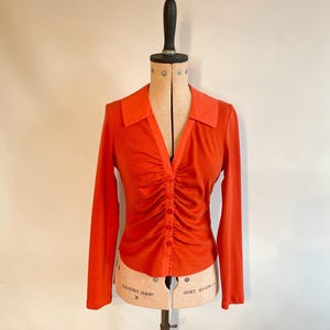 May include: An orange long-sleeve shirt with a V-neck and a button-down front. The shirt features a ruched design down the center and is displayed on a vintage dress form. The collar is a slightly lighter shade of orange.