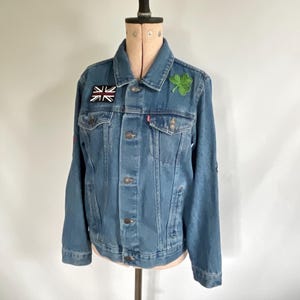 Op de afbeelding: Een lichtblauwe spijkerjas met knoopsluiting, twee borstzakken en zijzakken. Het heeft een Union Jack-patch en een groene klaverblad-patch. De jas wordt op een mannequin gepresenteerd.