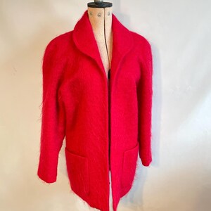 May include: A bright red, fuzzy, long-sleeved cardigan jacket featuring a wide collar, an open front design, and two large patch pockets on the lower front. The garment has a soft, textured, brushed appearance and is displayed on a mannequin.