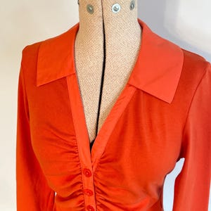 May include: Orange long-sleeved shirt with a contrasting orange collar. The shirt features a ruched front and three visible buttons. The garment is displayed on a beige dress form.