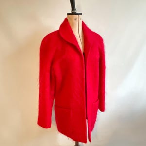 May include: A vibrant red coat with a classic collar and two front pockets. The coat is displayed on a vintage dress form, showcasing its tailored silhouette. The coat's color is a bold, eye-catching hue.