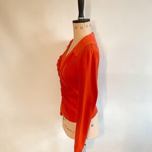May include: An orange long-sleeve top with a collared neckline and a ruched front. The top is displayed on a mannequin. The fabric appears to be a soft, lightweight material. The sleeves are long and fitted.