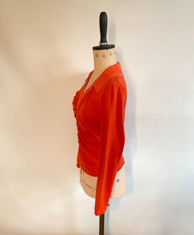 May include: An orange long-sleeve top with a collared neckline and a ruched front. The top is displayed on a mannequin. The fabric appears to be a soft, lightweight material. The sleeves are long and fitted.
