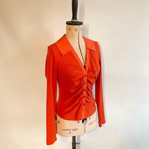 May include: An orange long-sleeved blouse with a collared neckline and a button-down front. The blouse features ruching down the center and is displayed on a vintage dress form. The dress form has a wooden top and a beige torso.
