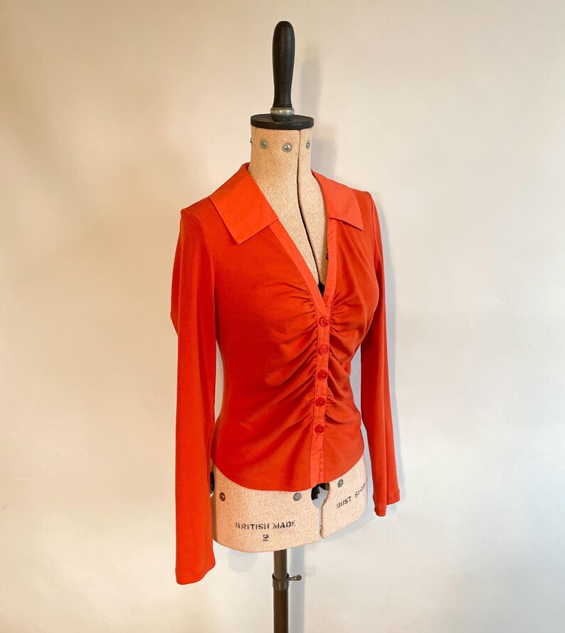 May include: An orange long-sleeved blouse with a collared neckline and a button-down front. The blouse features ruching down the center and is displayed on a vintage dress form. The dress form has a wooden top and a beige torso.
