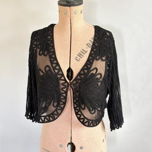 May include: Black bolero jacket with detailed floral embroidery. The jacket has a sheer, nude-coloured lining and three-quarter sleeves. The open front design highlights the intricate craftsmanship of the embroidered flowers.