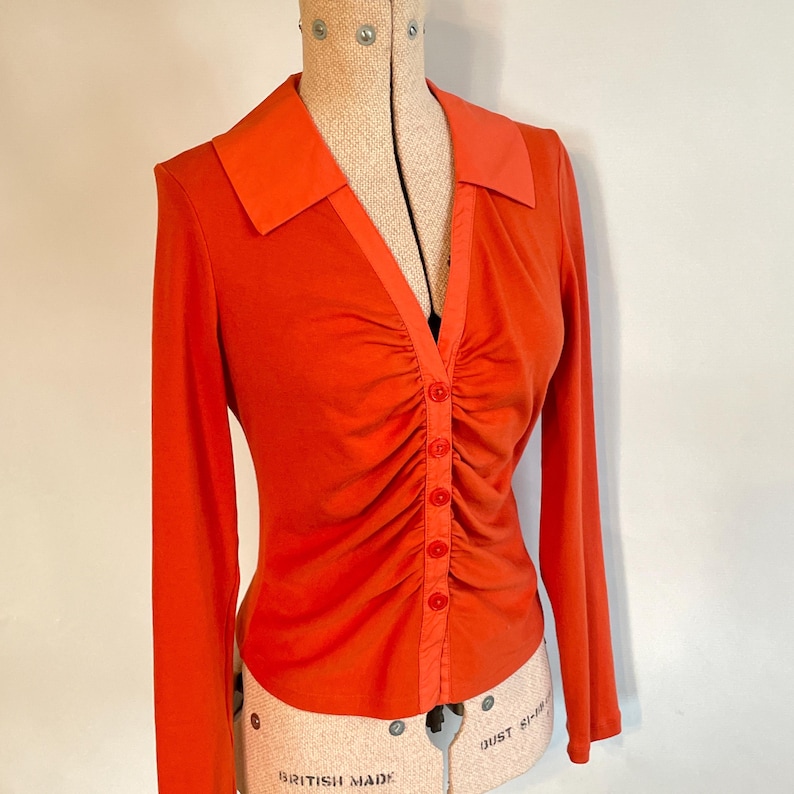 May include: An orange long-sleeve shirt with a collared neckline and a button-down front. The shirt features a ruched design down the center and is displayed on a vintage dress form. The collar is a lighter shade of orange.