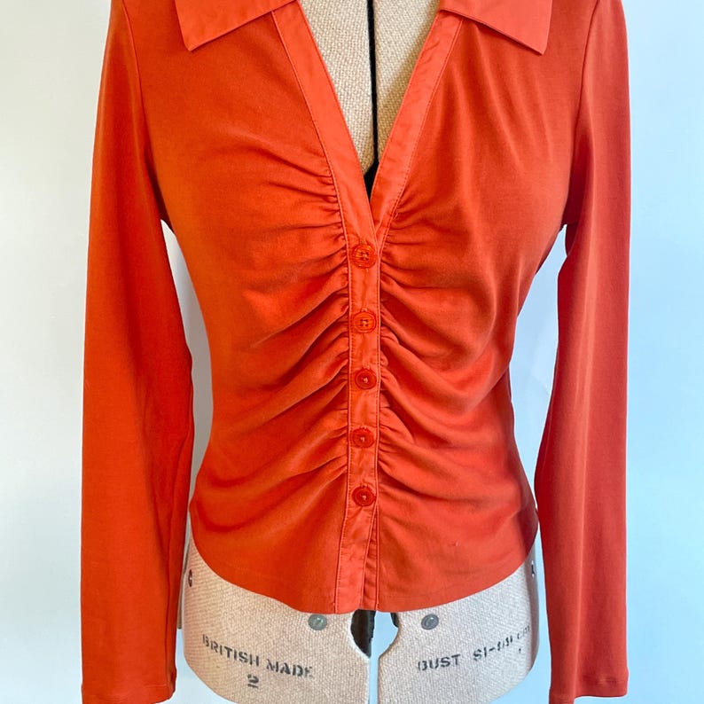 May include: An orange long-sleeve shirt with a collared neckline and a button-down front. The shirt features gathered fabric details down the center and is displayed on a dress form. The shirt is a solid color.