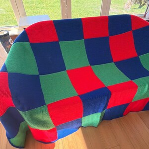 May include: A knitted blanket in a patchwork design, featuring squares of red, green, and blue. The blanket is draped over a sofa, showcasing its vibrant colors and geometric pattern. The blanket is made of a soft material.