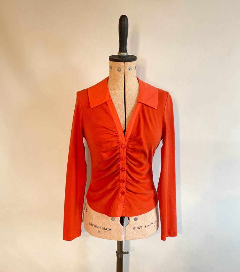 May include: An orange long-sleeve shirt with a V-neck and a button-down front. The shirt features a ruched design down the center and is displayed on a vintage dress form. The collar is a slightly lighter shade of orange.