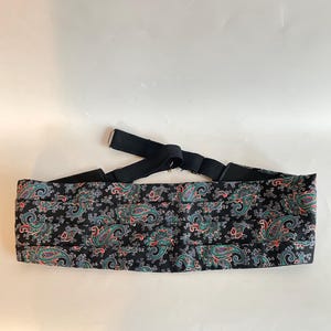 May include: A black cummerbund with a paisley pattern in teal, red, and gray. The cummerbund has a black elastic strap and a metal clasp. The accessory is designed to be worn around the waist.