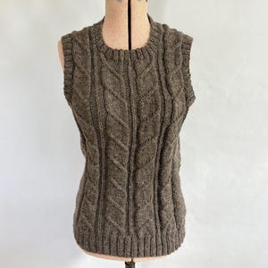 May include: A sleeveless brown knit sweater vest featuring a vertical cable knit pattern throughout the front. The garment has a round neckline and ribbed trim along the collar, armholes, and hem, constructed from a textured, heathered yarn.