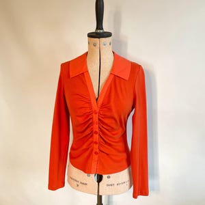 May include: An orange long-sleeve shirt with a ruched front and a collared neckline. The shirt has a button-down closure and is displayed on a vintage dress form. The color is a vibrant, solid orange.