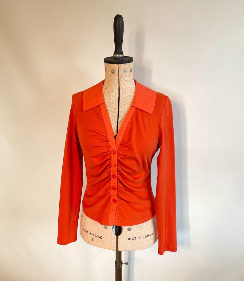 May include: An orange long-sleeve shirt with a ruched front and a collared neckline. The shirt has a button-down closure and is displayed on a vintage dress form. The color is a vibrant, solid orange.