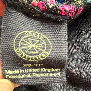 Może przedstawiać: Zbliżenie czarnej metki z tkaniny z logo Urban Outfitters w ż&oacute;łtym okręgu. Na metce widnieje napis "Made in United Kingdom Fabriqu&eacute; au Royaume-uni" i rozmiar "XS-TP".
