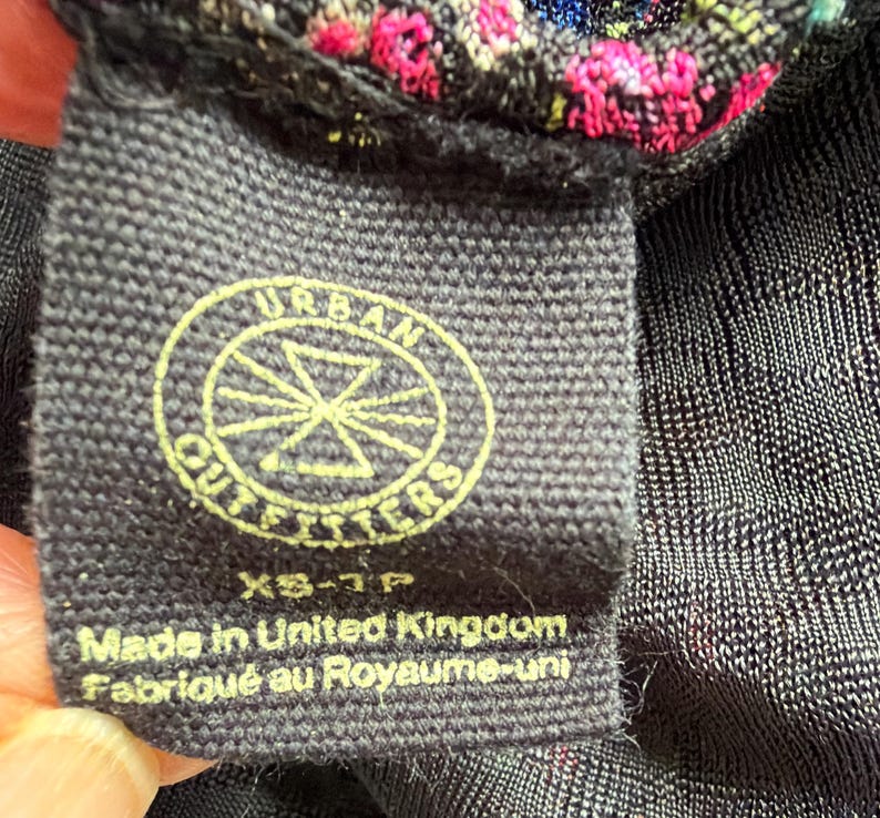 Może przedstawiać: Zbliżenie czarnej metki z tkaniny z logo Urban Outfitters w ż&oacute;łtym okręgu. Na metce widnieje napis "Made in United Kingdom Fabriqu&eacute; au Royaume-uni" i rozmiar "XS-TP".