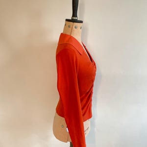 May include: A burnt orange long-sleeve top with a collared neckline and a ruched design on the front. The top is displayed on a mannequin, showcasing its tailored fit and style. The fabric appears to be a soft knit material.