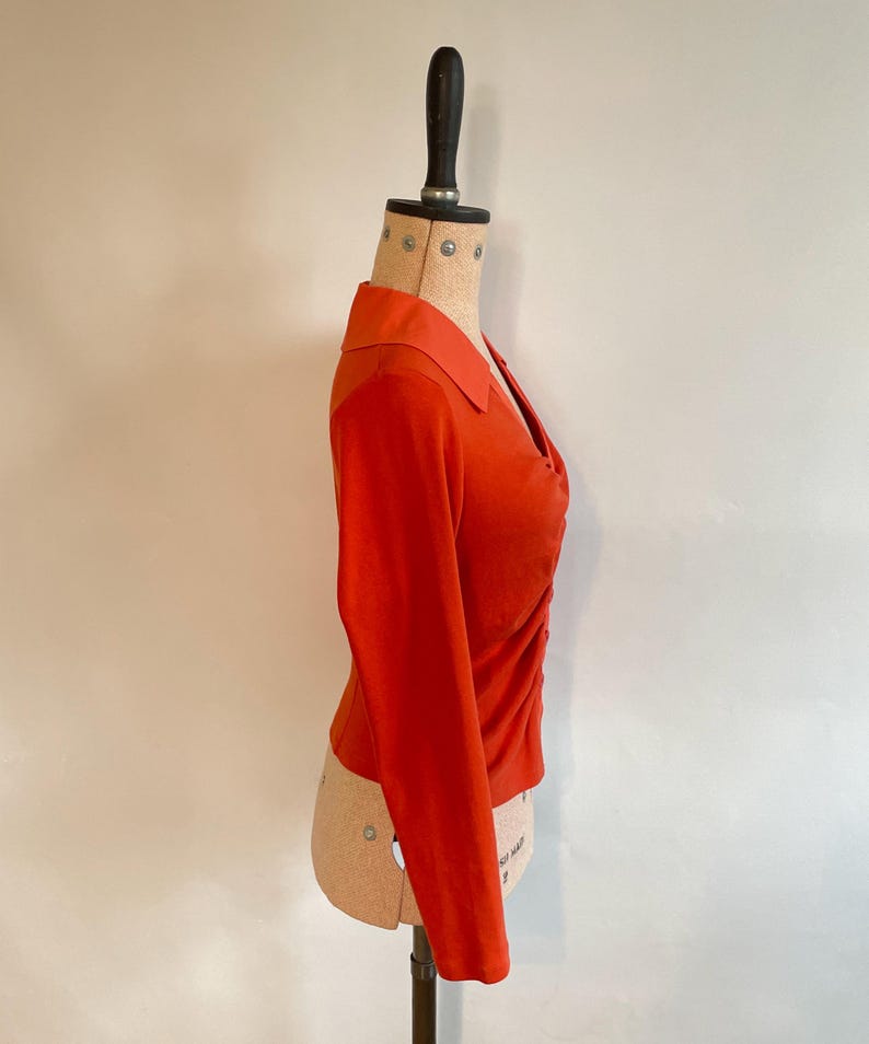 May include: A burnt orange long-sleeve top with a collared neckline and a ruched design on the front. The top is displayed on a mannequin, showcasing its tailored fit and style. The fabric appears to be a soft knit material.