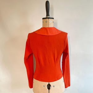 May include: A bright orange long-sleeved top with a wide, rounded collar. The fitted bodice is displayed on a vintage dress form. The top's color is a vibrant, solid hue, and the sleeves are long and fitted. The dress form is beige and has a black neck piece.