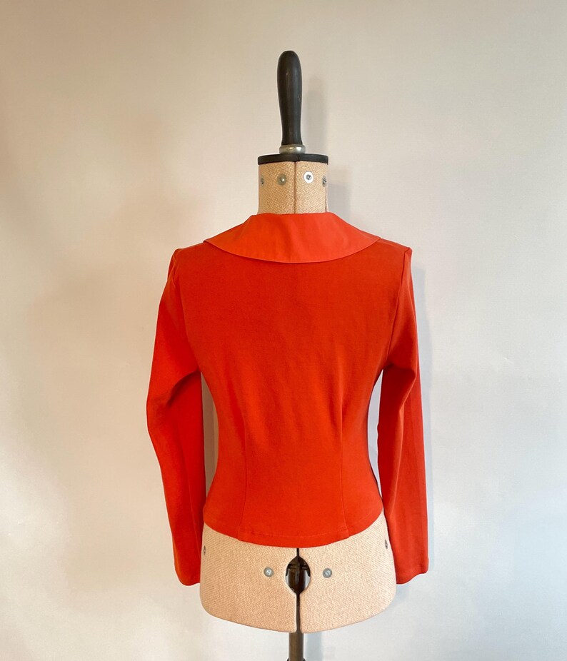 May include: A bright orange long-sleeved top with a wide, rounded collar. The fitted bodice is displayed on a vintage dress form. The top's color is a vibrant, solid hue, and the sleeves are long and fitted. The dress form is beige and has a black neck piece.