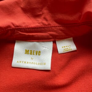 May include: Close-up of a vibrant orange shirt with a collar. The label reads "Maeve by Anthropologie" and "Small Made in Cambodia". The fabric appears to be a soft, textured material.