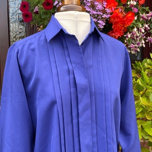 May include: A vibrant royal blue shirt with vertical pleats down the front. The long-sleeved shirt has a classic collar and is displayed on a mannequin. The background features colorful flowers.