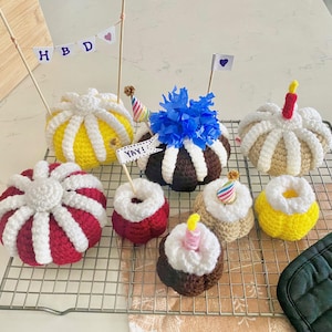 Könnte beinhalten: Eine Sammlung handgefertigter gehäkelter Geburtstagskuchen in verschiedenen Farben, darunter Gelb, Rot und Braun. Einige Kuchen haben dekorative Topper wie Kerzen und Fähnchen mit dem Text "HBD" und "YAY!"