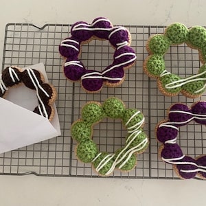 Puede incluir: Cinco donuts de crochet en varios colores, incluyendo morado, verde y marrón, con glaseado blanco. Los donuts están dispuestos en una rejilla de alambre.