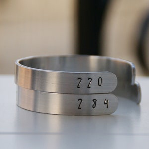 Könnte beinhalten: Ein silbernes Armband mit zwei Bändern. Das obere Band hat die Zahlen "220" eingraviert. Das untere Band hat die Zahlen "284" eingraviert.